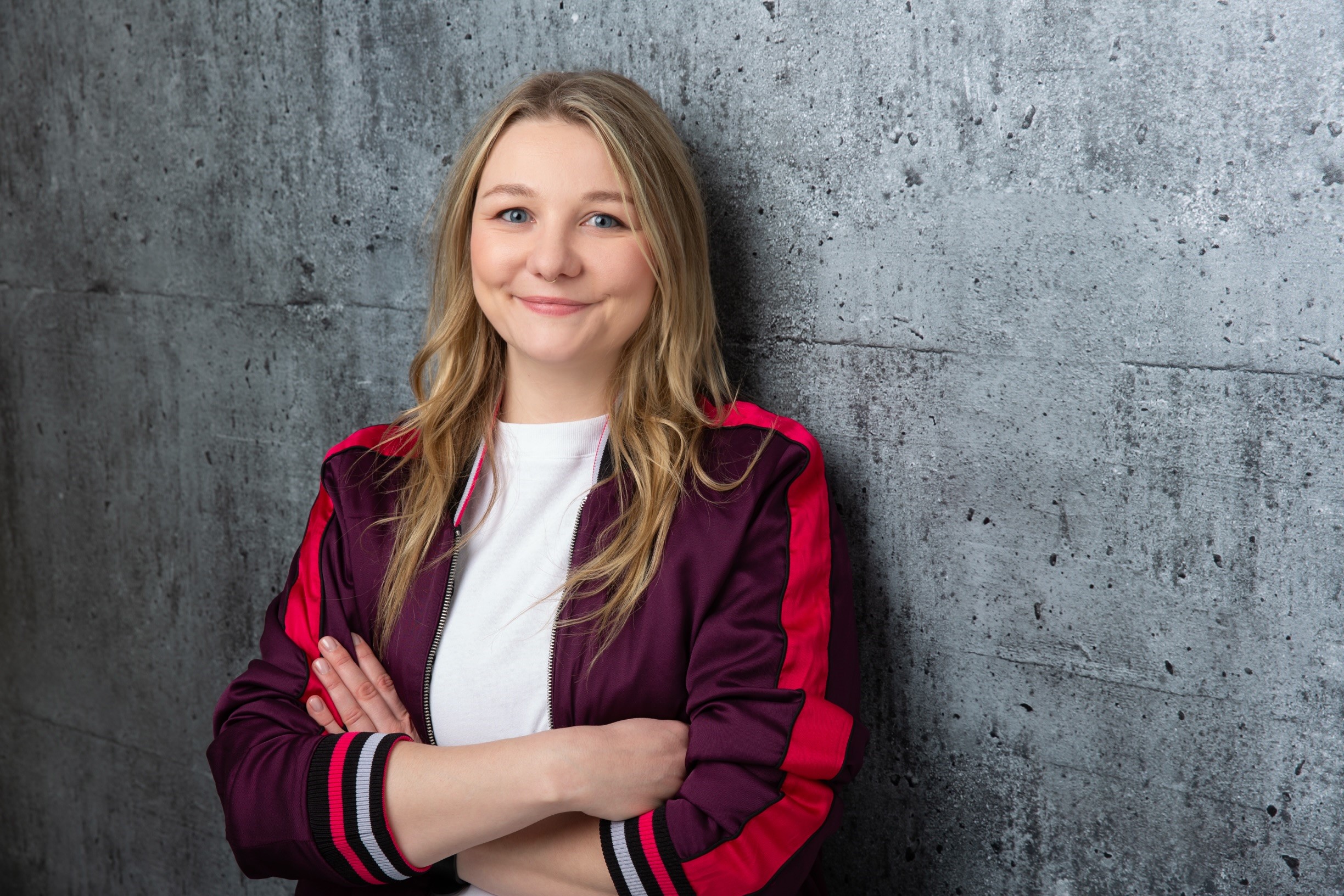 Foto von Johanna Niedergesäß mit ila-roter Jacke und deutlich lächelnd vor einer grauen Wand. (Quelle: privates Foto.) Foto von Johanna Niedergesäß mit ila-roter Jacke und deutlich lächelnd vor einer grauen Wand.